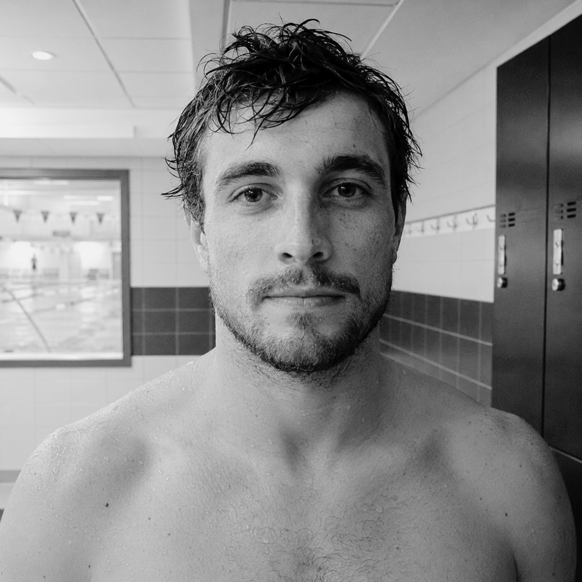 Male swimmer looking directly at camera in a leisure centre changing room after training, damp skin, calm expression